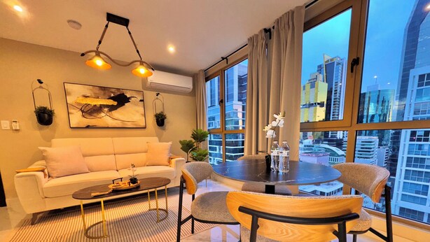 Una sala de estar moderna con sofá, mesa de centro y sillas, una mesa de comedor con botellas de agua, y grandes ventanales con vista a la ciudad.