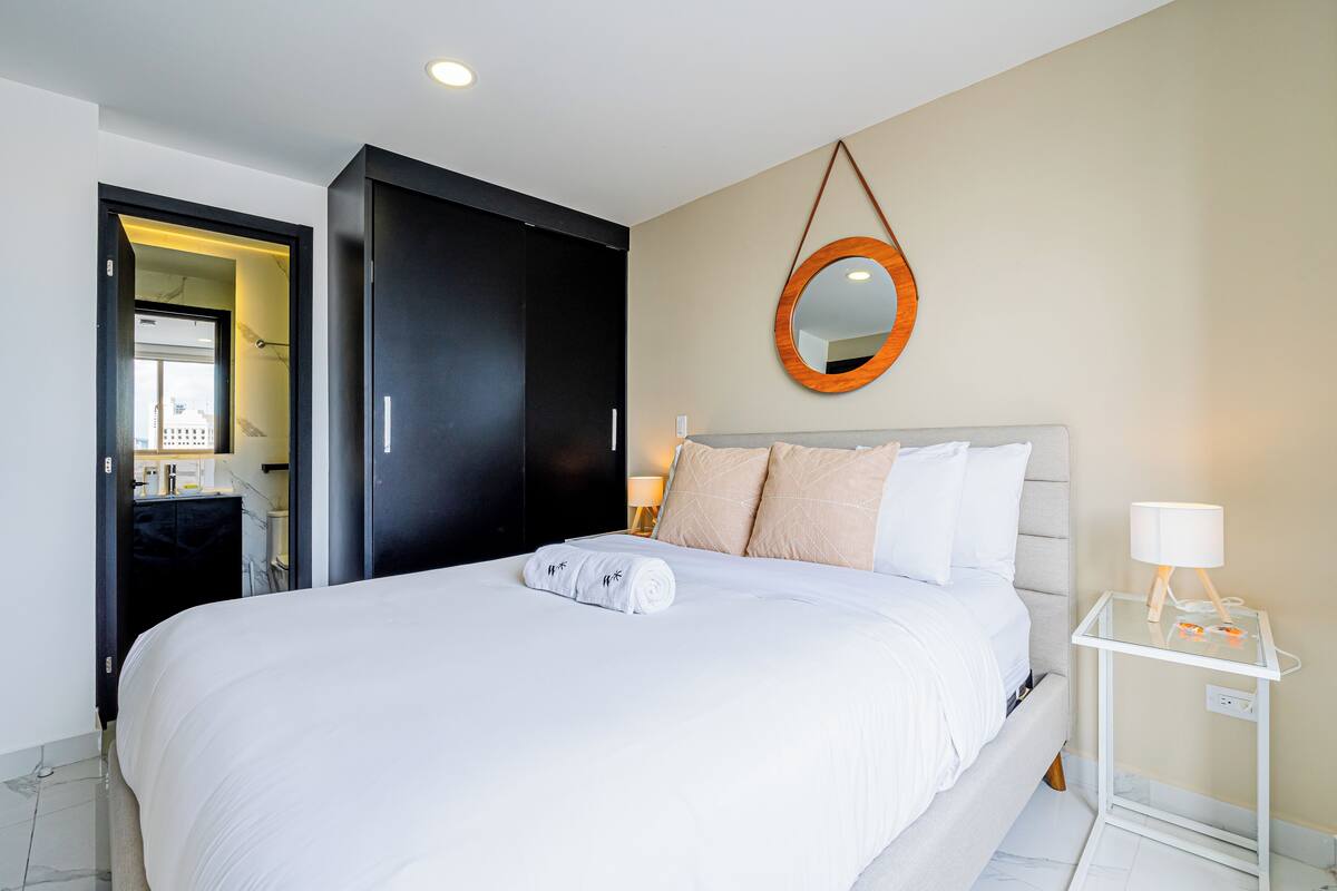 A neatly made bed with a rolled towel, a bedside table with a lamp, and a mirror on the wall.