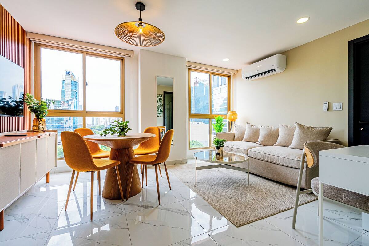 A modern living room with a round dining table, orange chairs, a beige sofa, and a glass coffee table.