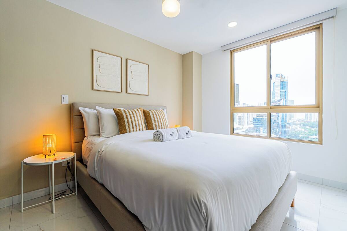 A modern hotel room with a large bed, a bedside table with a lamp, and a city view through a large window.