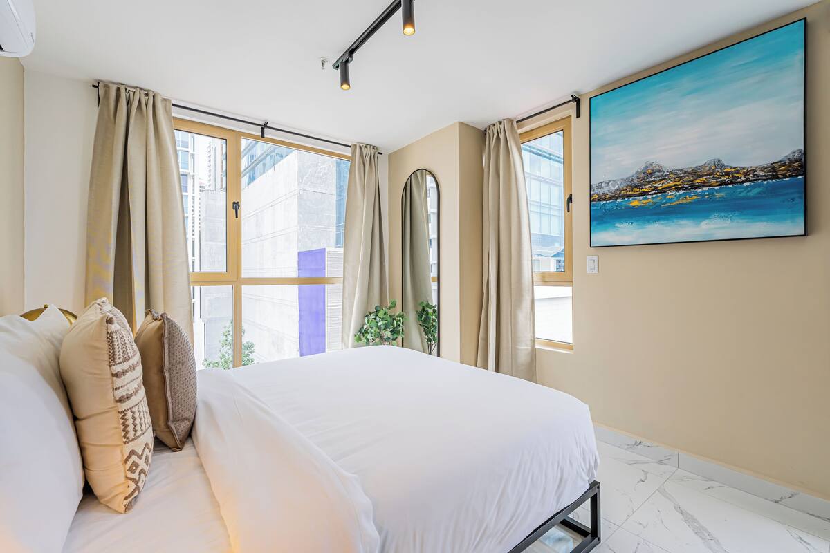 A bedroom with a large bed, beige curtains, a painting of a coastal scene, and a window with a view of buildings.