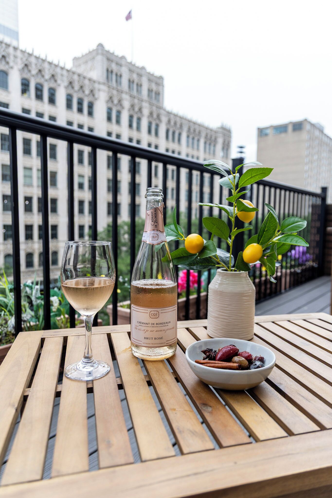 A table with a bottle of Brut Rose wine, a glass of wine, and a bowl of dried fruits on a balcony with a city view.