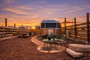 Une caravane Airstream ancienne, avec une table de pique-nique et des chaises, installée sur un terrain recouvert de gravier, entourée de clôtures en bois, et avec un coucher de soleil en arrière-plan.