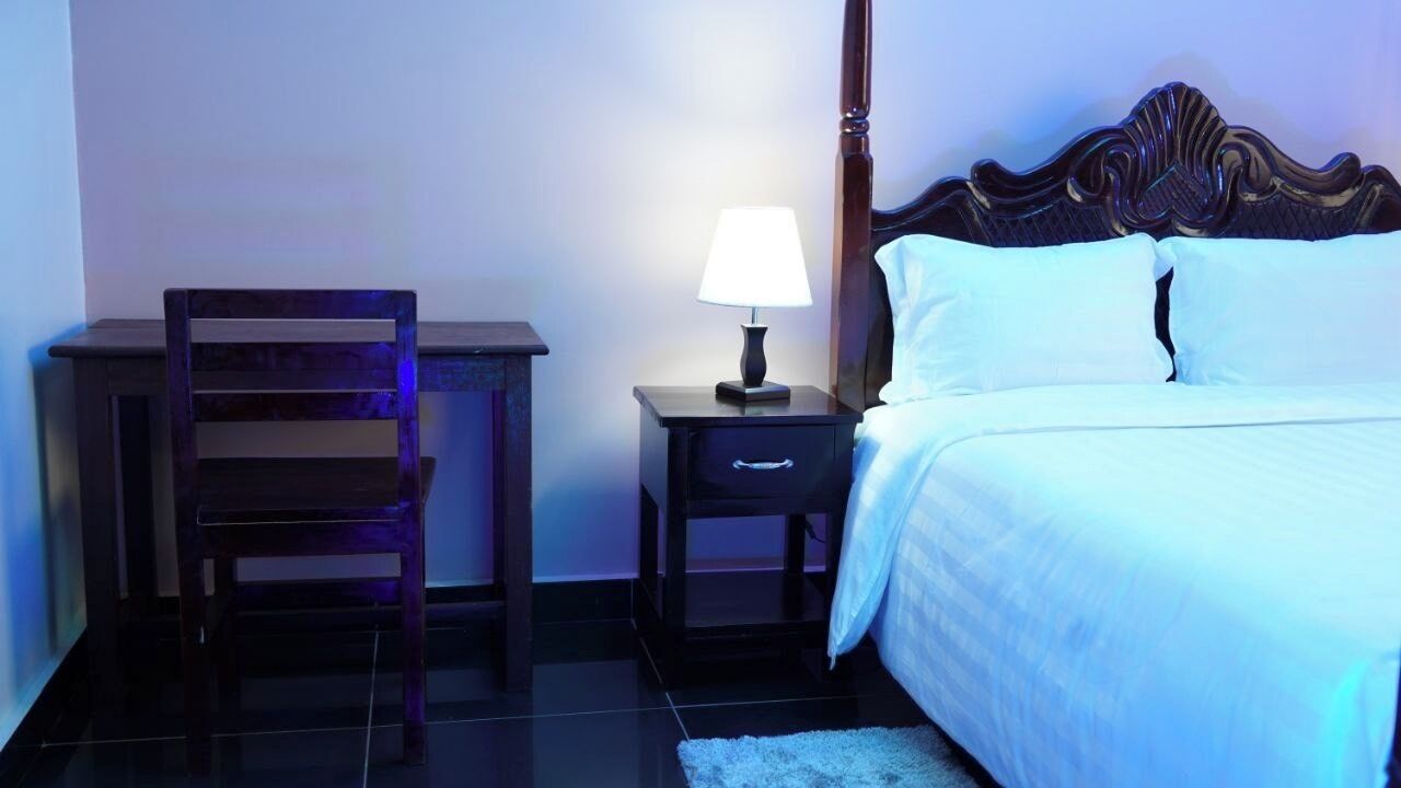 A hotel room with a bed, a nightstand with a lamp, a wooden chair, and a small desk.