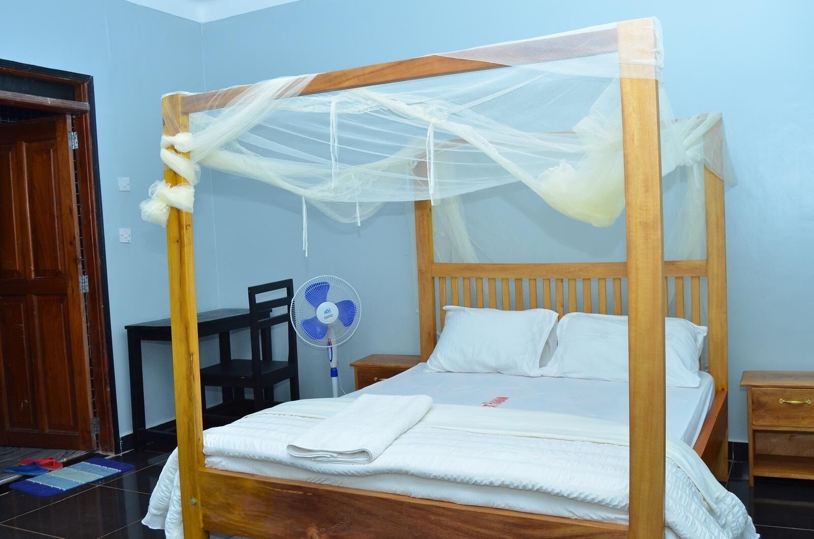 A single bed with a wooden frame and a mosquito net, a bedside table, a chair, and a standing fan.