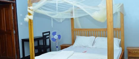 A single bed with a wooden frame and a mosquito net, a bedside table, a chair, and a standing fan.
