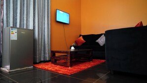 A modern living room with a flat-screen TV mounted on the wall, a refrigerator, a wooden coffee table, and a black sofa with orange cushions. - Kamfart Safari Lodge (Jinja)