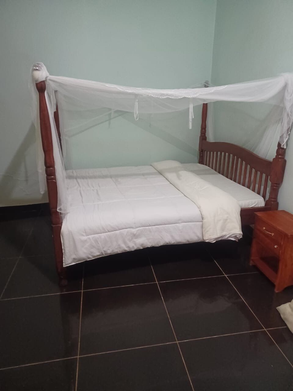 A bed with a wooden frame and a white bedspread, a small wooden nightstand, and a tiled floor.