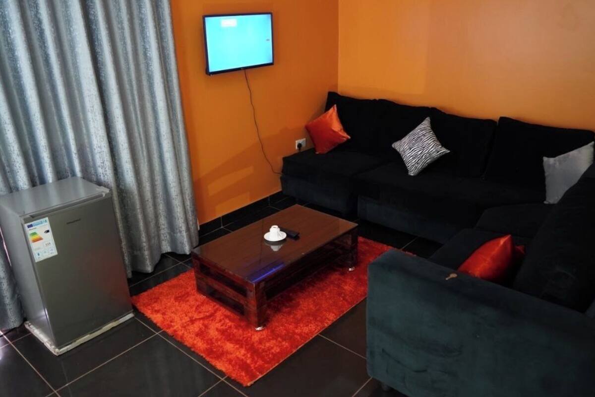 Un salon avec un canapé noir, un tapis rouge, une petite table sur laquelle se trouve une théière, un réfrigérateur gris et un téléviseur fixé au mur.