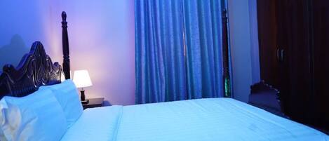 A neatly made bed with white linens, a wooden headboard, a nightstand with a lamp, and a window with blue curtains.