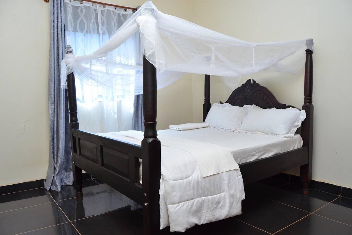 Un lit à baldaquin avec un cadre en bois foncé, des draps blancs et un ciel de lit transparent.