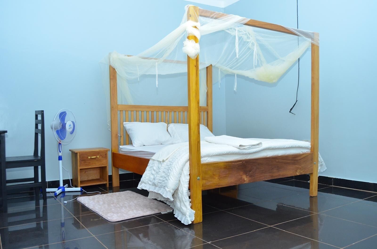 Uma cama de solteiro com dossel, mesa de cabeceira, cadeira e ventoinha em um quarto com piso de azulejos e paredes azul-claras.