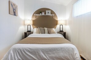 En pænt redt seng med stor, rund vægdekoration over, to sengeborde med lamper, og en lille hylde med en plante og et billedramme.