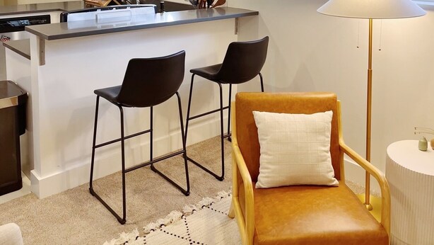 Une cuisine moderne dotée d’un îlot central, de deux tabourets de bar, d’une chaise en bois avec un coussin et d’un lampadaire.