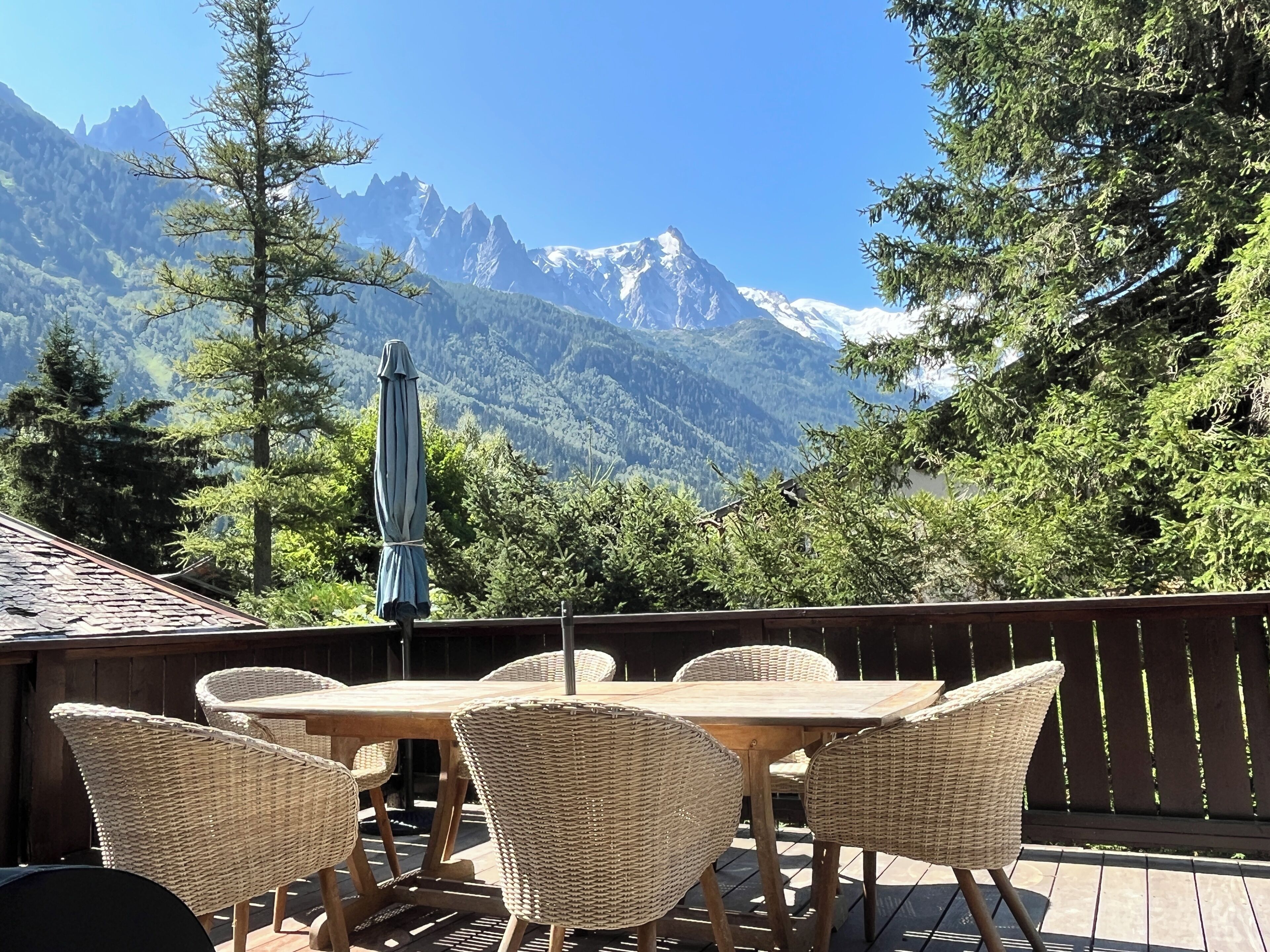 A wooden deck with wicker furniture and a table, overlooking a mountainous landscape with snow-capped peaks.
