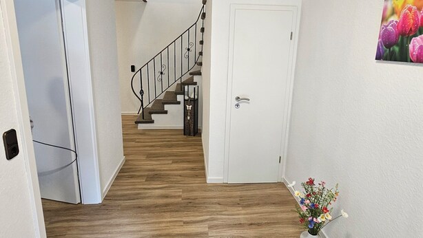 A hallway with wooden flooring, a white door, a staircase with a black railing, and a painting of flowers on the wall. - Bright vacation apartment in East Frisia with large garden and terrace (Friedeburg)