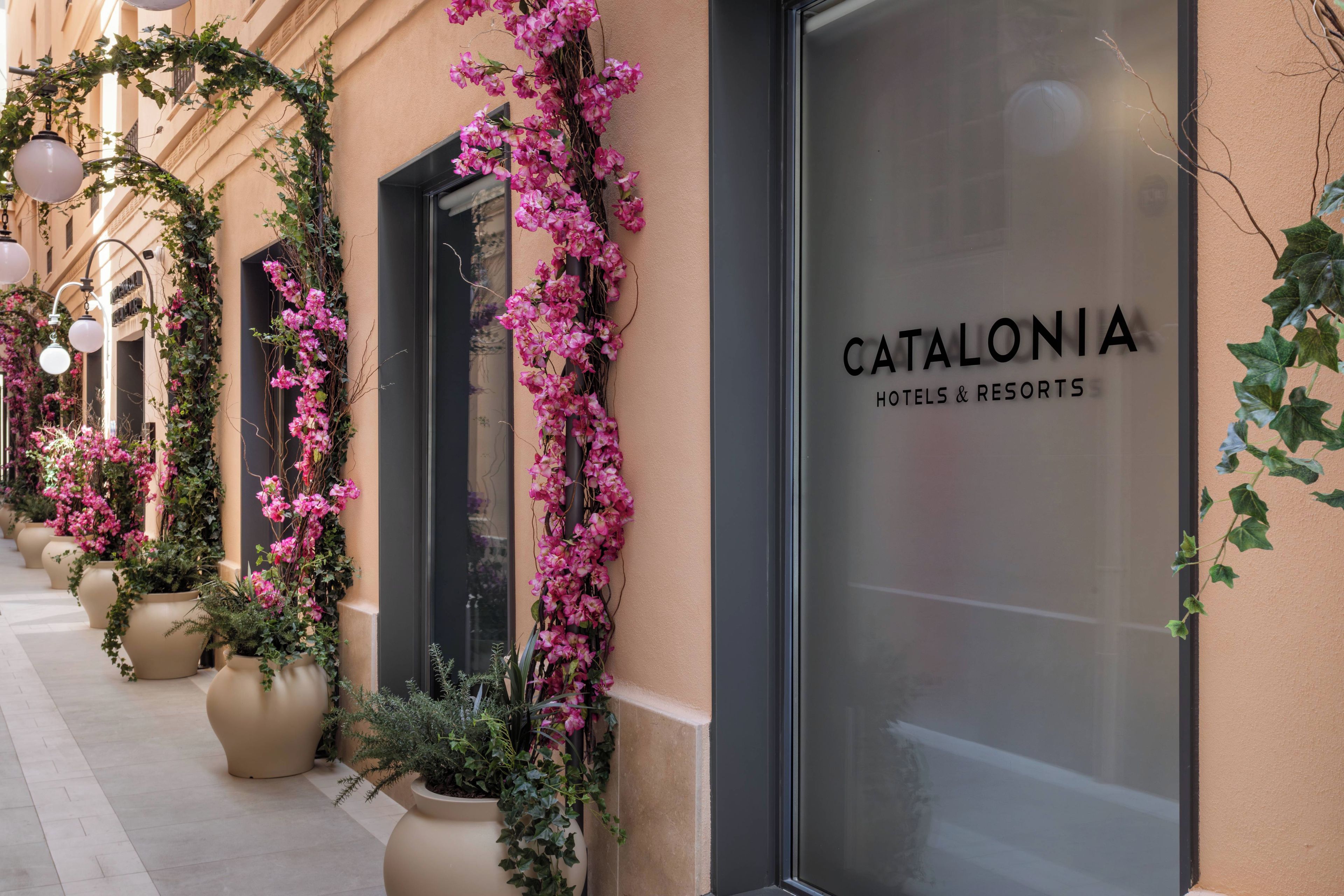 Entrada de hotel con fachada de vidrio, flores rosadas y plantas en macetas.