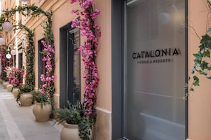 Entrada de hotel con fachada de vidrio, flores rosadas y plantas en macetas.