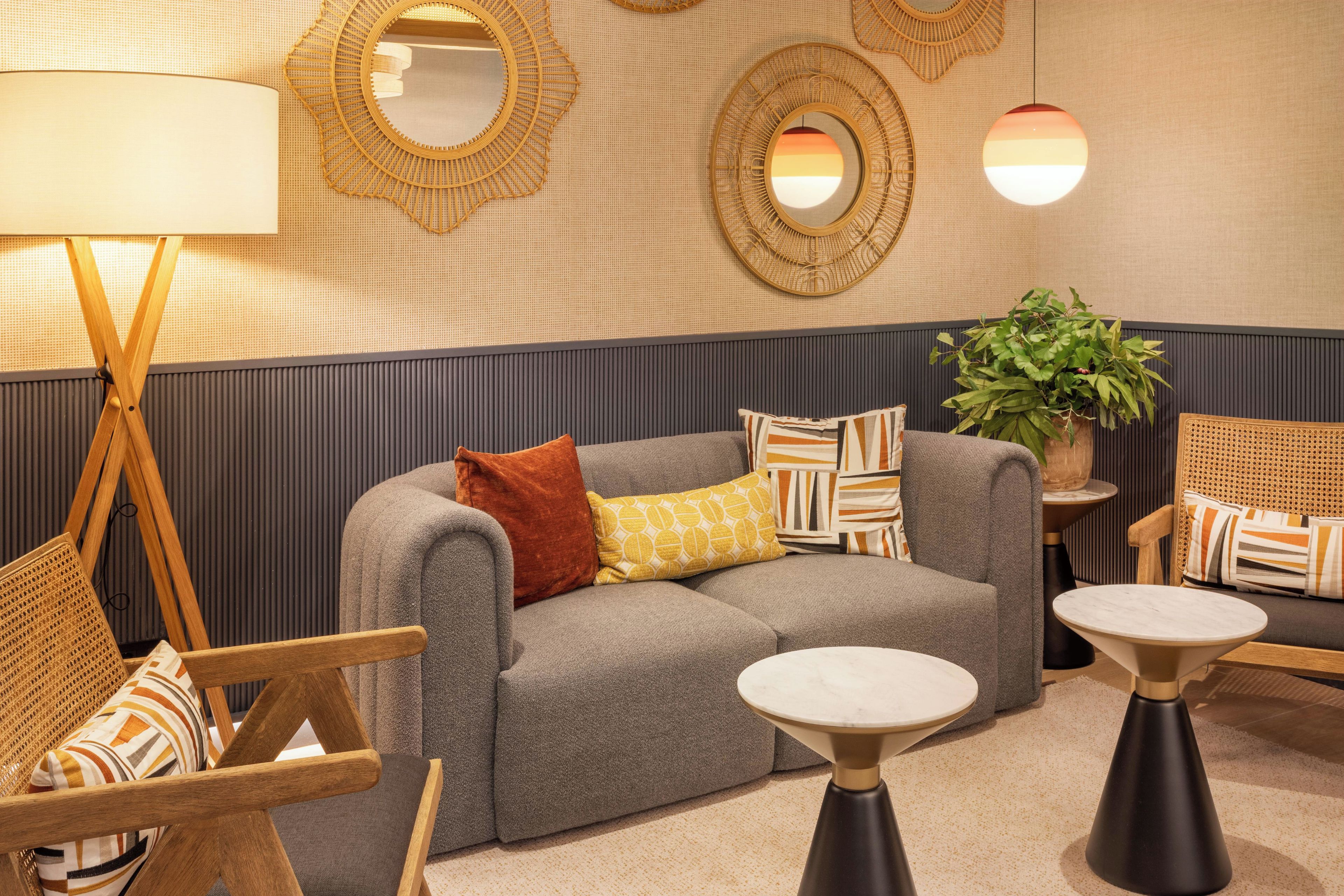 Un área de descanso moderna con un sofá gris, cojines estampados, una silla de madera, una mesita y espejos decorativos en la pared.