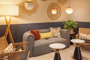 Un área de descanso moderna con un sofá gris, cojines estampados, una silla de madera, una mesita y espejos decorativos en la pared.