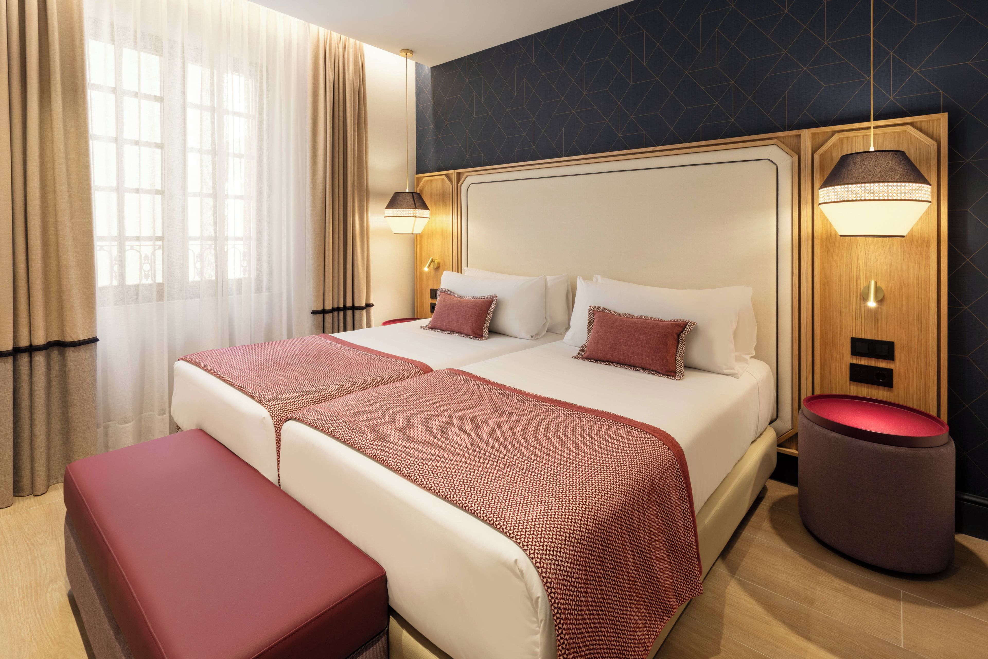 A double bed with a patterned bedspread, two bedside lamps, a wooden headboard, and a red bench.