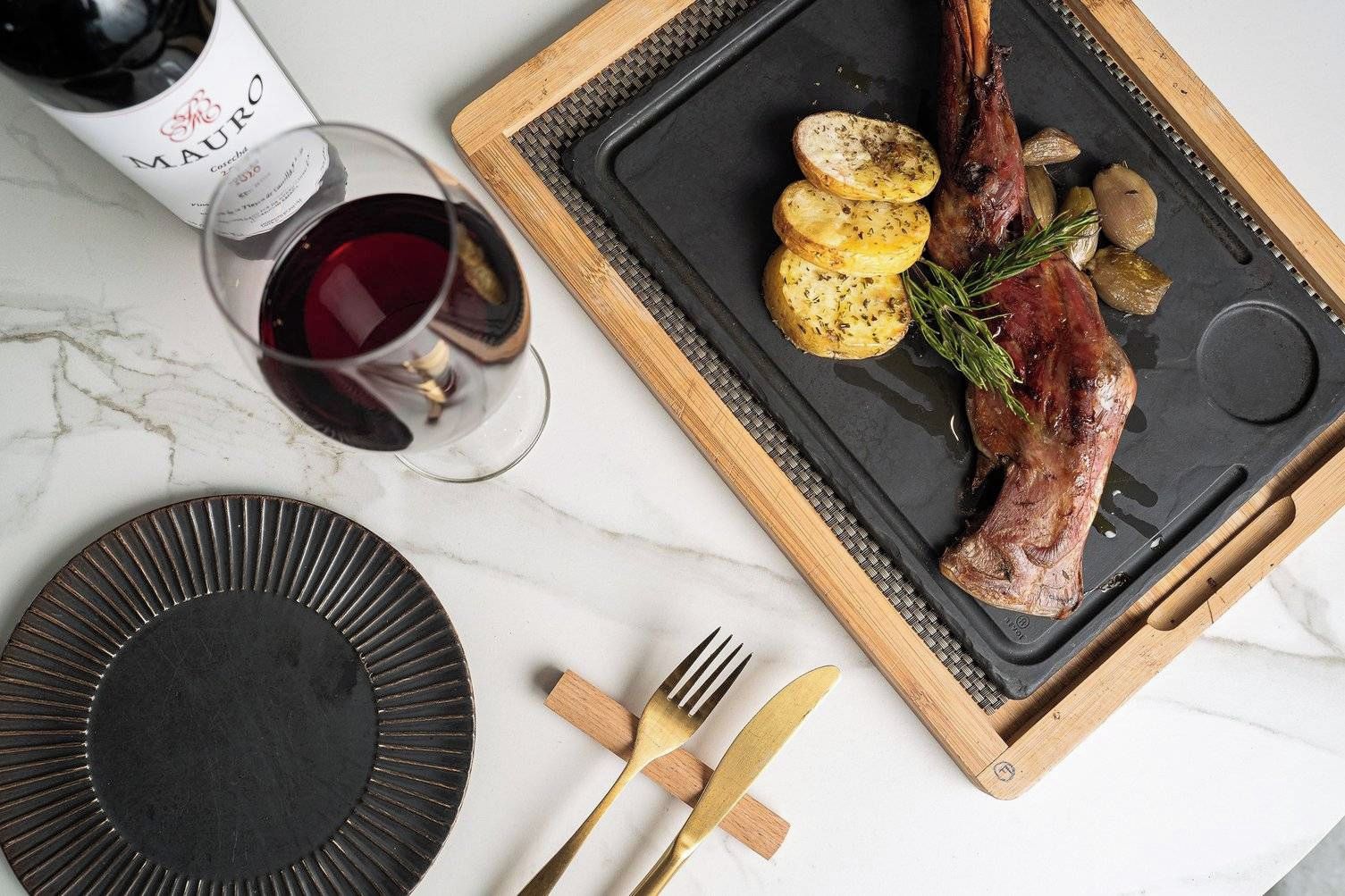 Una mesa de comedor con un plato de carne asada, papas y hierbas, acompañado de una copa de vino tinto y cubiertos sobre una superficie de mármol.