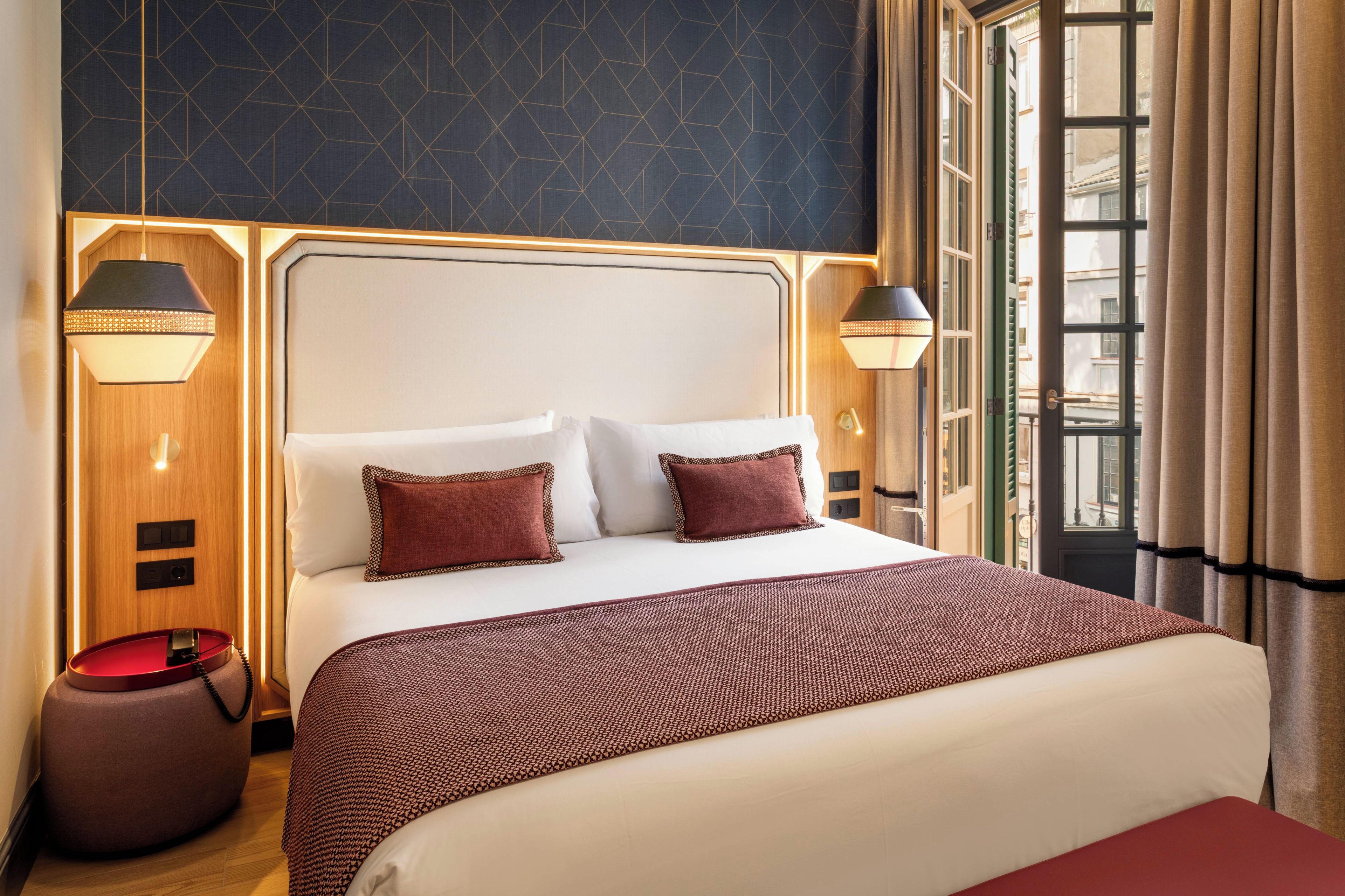 Une chambre d’hôtel avec un grand lit, deux lampes de chevet, un téléphone rouge et une fenêtre avec des rideaux.