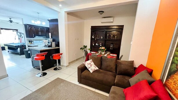 A modern living room with a brown sofa, red cushions, and a red bar stool. A dining table with a red tablecloth and a vase of flowers is visible. The room has a large window, a ceiling fan, and a kitchen area with dark cabinets and a white countertop.