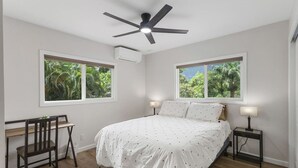 A bedroom with a bed, two bedside tables with lamps, a desk with a chair, two windows with views of greenery, a ceiling fan, and an air conditioning unit. - Secluded Forest Retreat Full Kitchen, AC, W/D (Kailua)