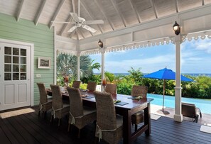 A covered outdoor dining area with wicker chairs and a table, overlooking a pool and lush greenery.