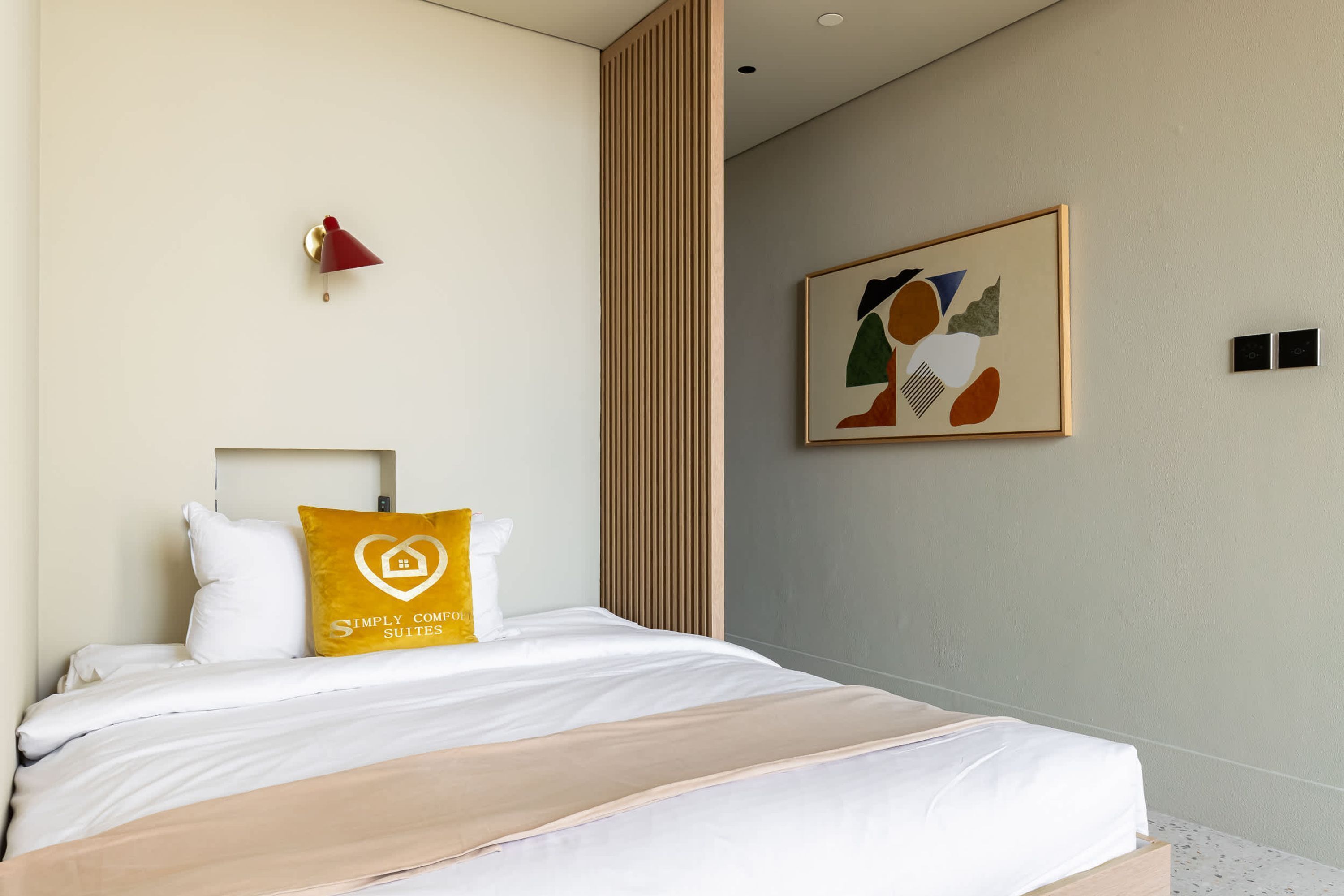 Une chambre d’hôtel moderne avec un lit soigneusement fait, une lampe murale rouge, un tableau abstrait encadré et un panneau en bois sur le mur.