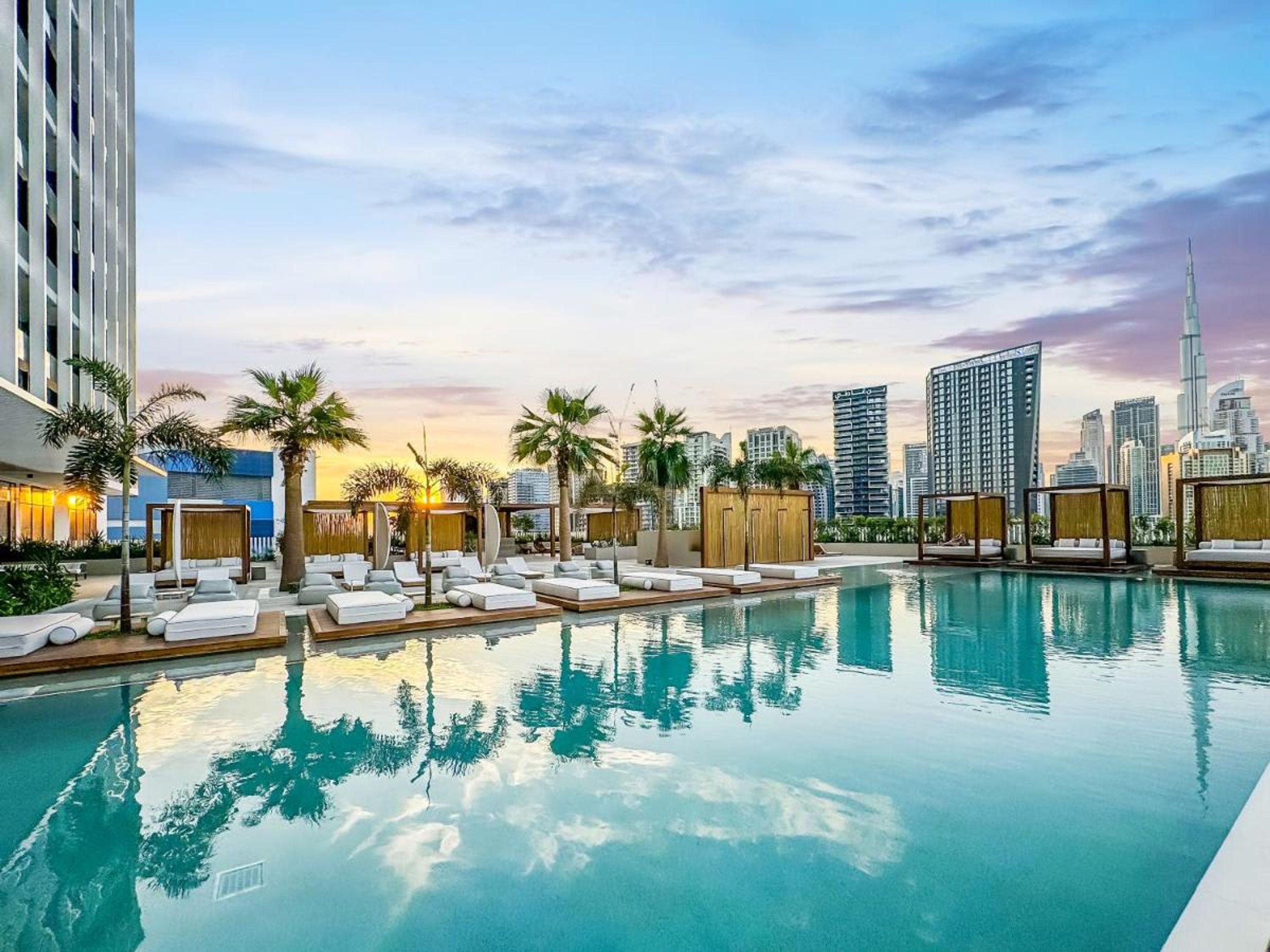 A rooftop pool with lounge chairs and palm trees, surrounded by high-rise buildings and a sunset sky.