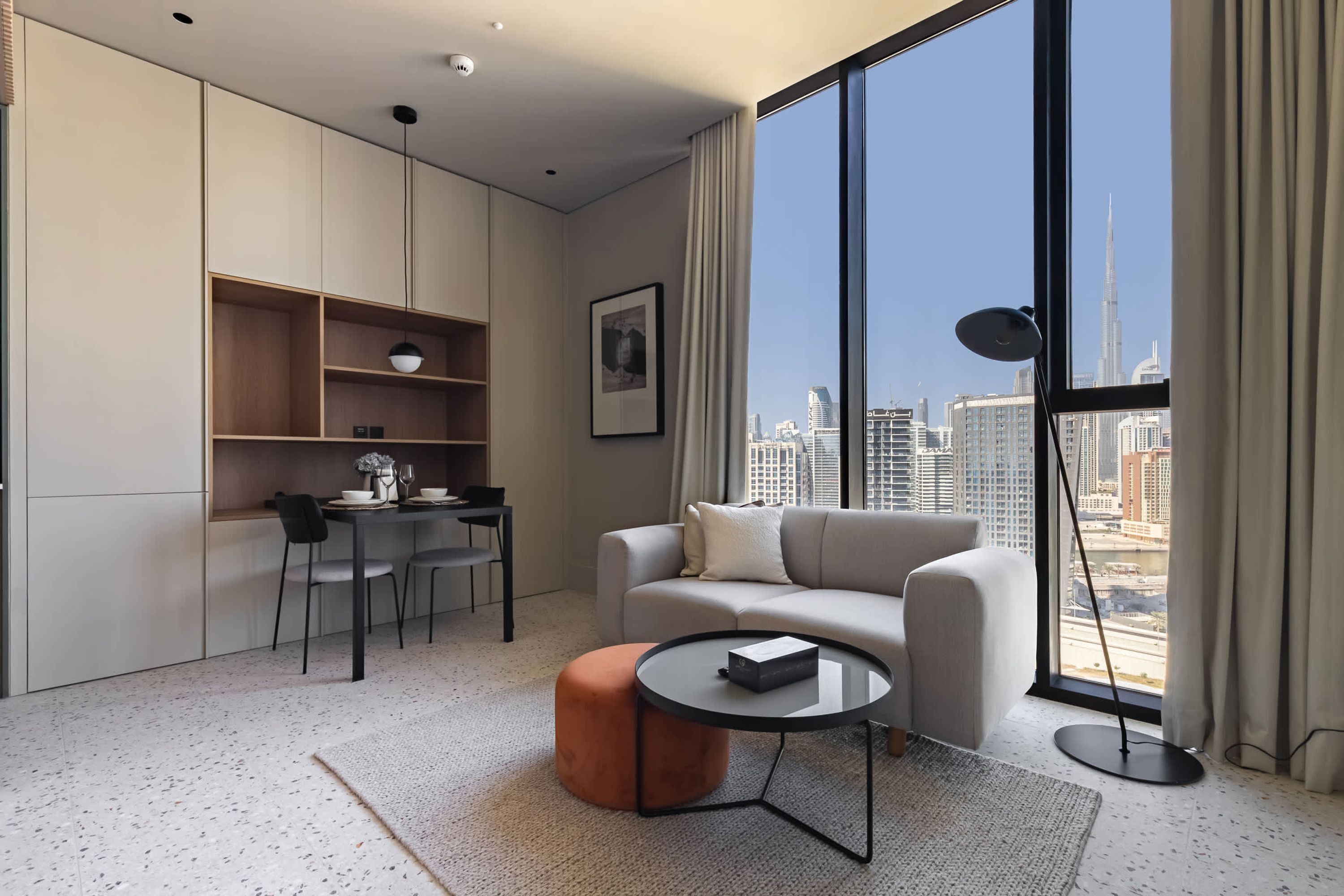 A modern living room with a sofa, coffee table, and dining area, featuring a city view through large windows.