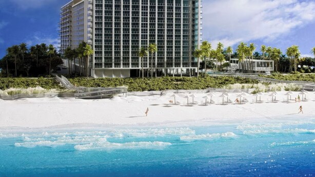 Ein Strandhotel mit einem großen Gebäude, Palmen und einem Sandstrand mit Sonnenschirmen.