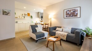 A modern living room with a grey sofa, a white armchair, and a wooden coffee table. A kitchen is visible in the background with white cabinets and a dining area. - Brisa do Mar in S o Martinho do Porto (Alcobaça)