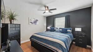 Un dormitorio con cama, mesitas de noche, ventilador de techo, ventana con cortinas, escritorio con silla y planta en maceta.