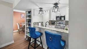 Una cocina moderna con armarios blancos, encimera de granito y horno empotrado. Hay dos taburetes azules y luces colgantes sobre la encimera.