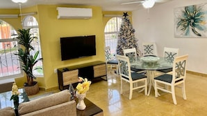 A living room with a flat-screen TV, a glass dining table with chairs, a Christmas tree, and a ceiling fan.
