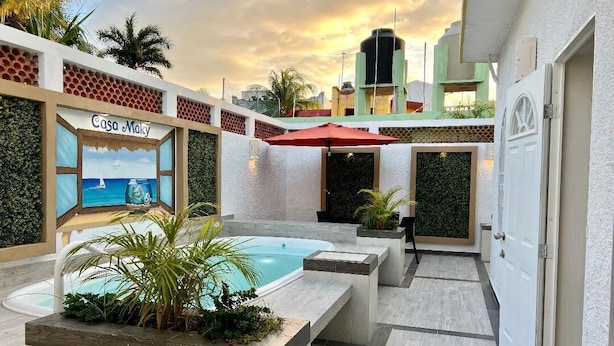 A pool area with a sign reading "Casa Maky," surrounded by a white wall with a patterned design, a red umbrella, and a door to the right.