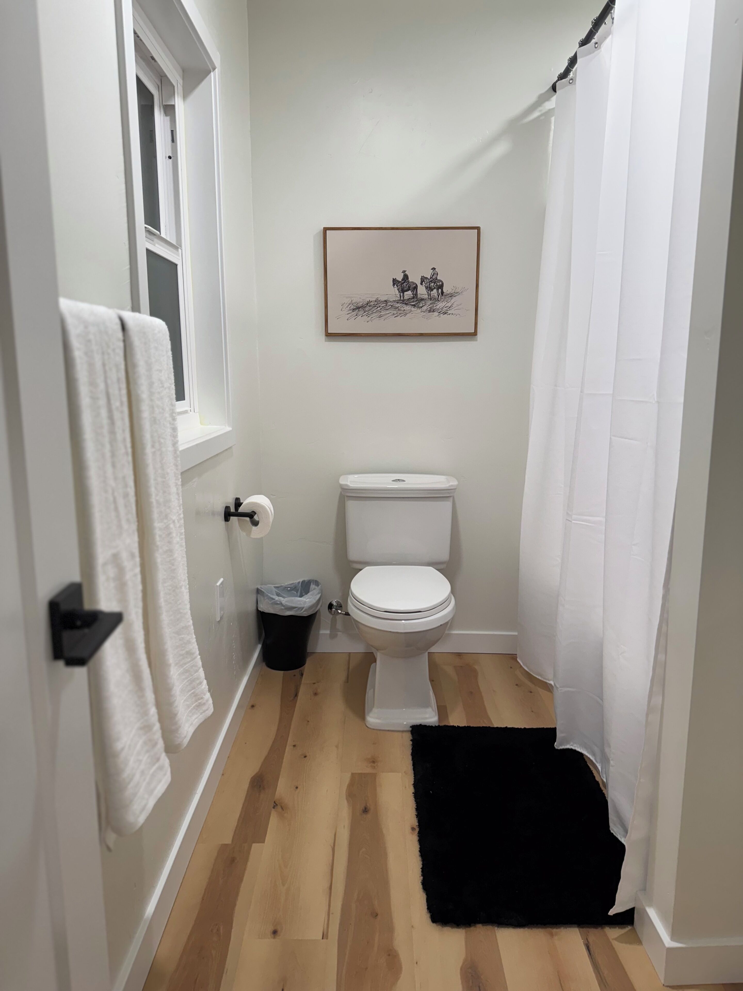 A small bathroom with a toilet, a black rug, a white towel on a rack, a trash bin, and a framed picture on the wall.