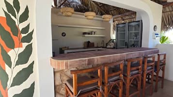 Een bar met een stenen aanrecht en houten stoelen, een keuken zichtbaar door een boog, en een rieten dak erboven.