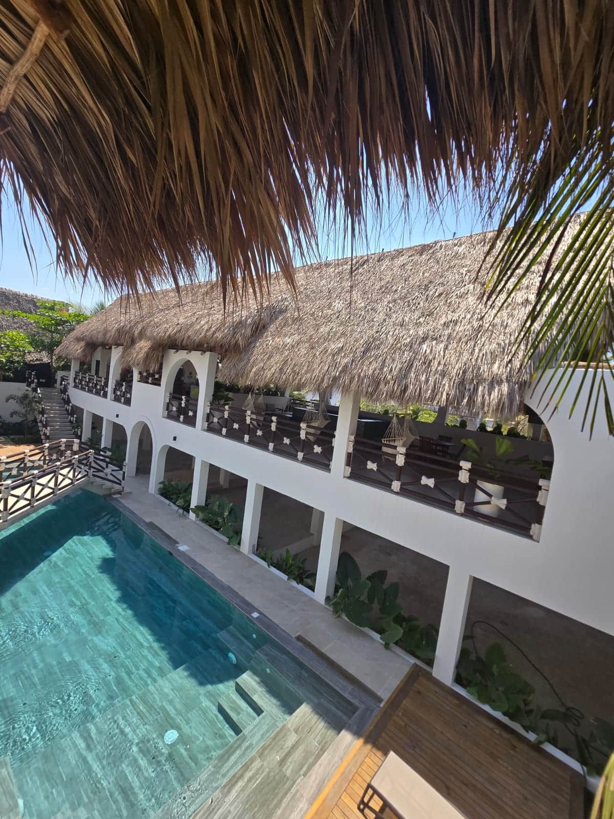 Un espace aménagé au bord de la piscine, doté d’une structure à toit de chaume, d’une piscine à eau claire et d’un escalier menant à un niveau supérieur.