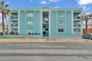 Un immeuble de couleur bleu clair, à plusieurs étages, doté de balcons, d’une entrée centrale et d’un parking situé à l’avant.