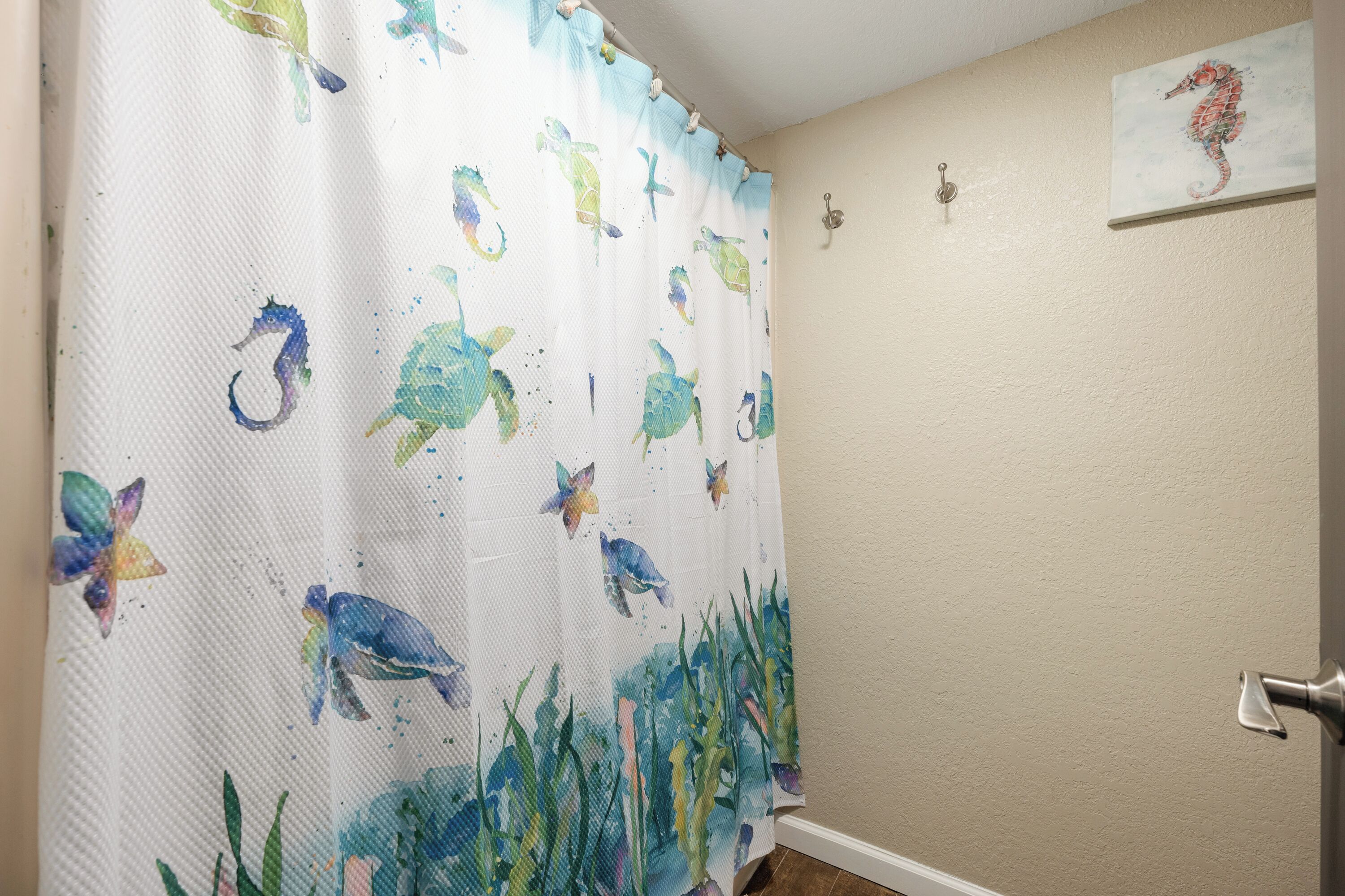 A bathroom with a shower curtain featuring sea creatures, a wall-mounted light fixture, and a seahorse painting.