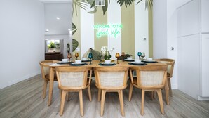 A dining area with a wooden table and wicker chairs, a tropical-themed wall mural, and a view of a living room in the background. - NEW! Serene Designer 3BR near Polo & Tennis- SaltPool+Spa, GameRm, Firepit, Golf (Indio)