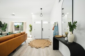 A modern living room with a sofa, a round woven rug, a black console table with a vase and plants, and a large mirror. - NEW! Serene Designer 3BR near Polo & Tennis- SaltPool+Spa, GameRm, Firepit, Golf (Indio)
