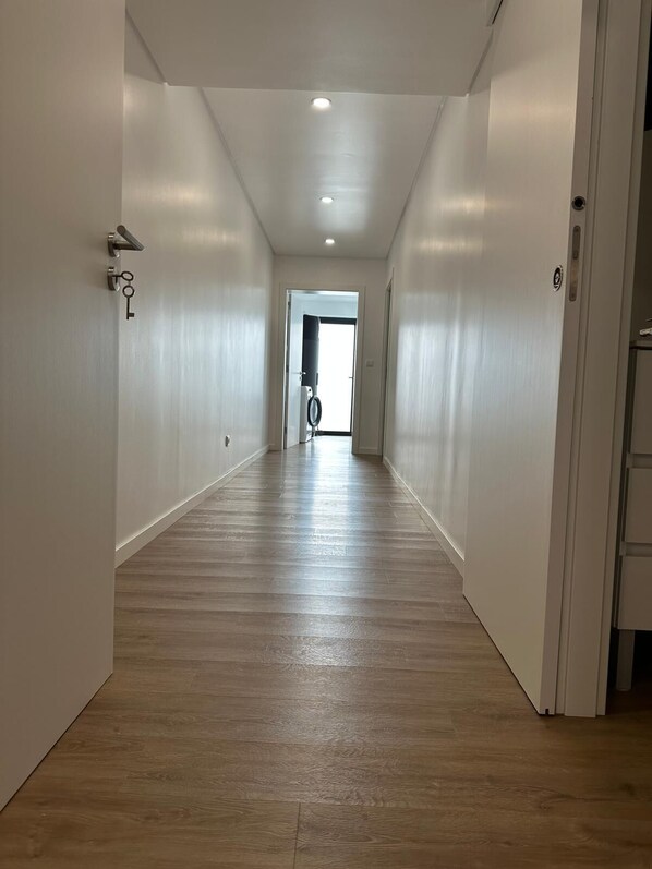 Un couloir étroit et bien éclairé, doté d’un plancher en bois et de murs blancs, qui mène à une porte avec un trou de serrure et une poignée.