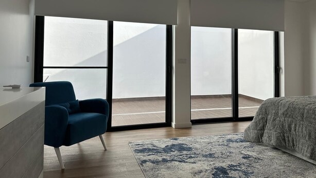 Ein modernes Schlafzimmer mit großen Schiebeglasfenstern, die auf einen Balkon führen, einem blauen Sessel, einem Bett mit einer gemusterten Decke und einem Teppich.