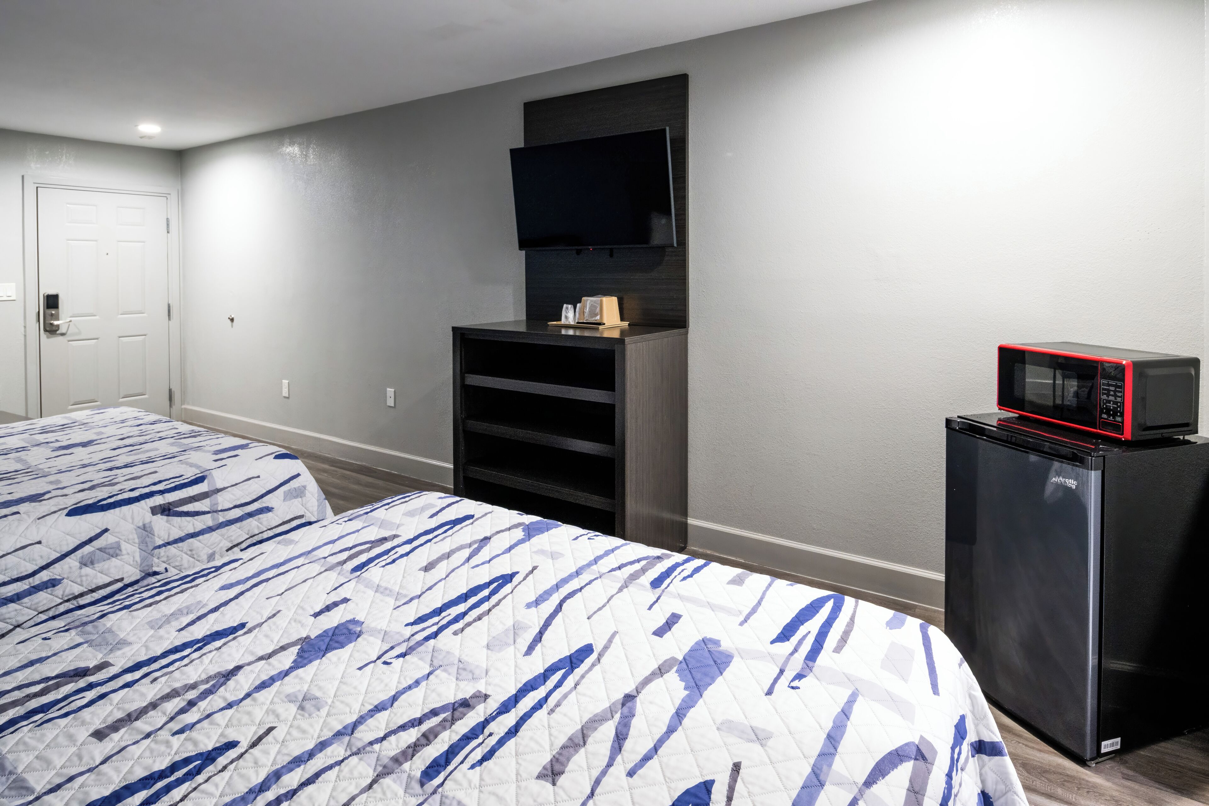A hotel room with a bed, a TV mounted on the wall, a small refrigerator, and a door.