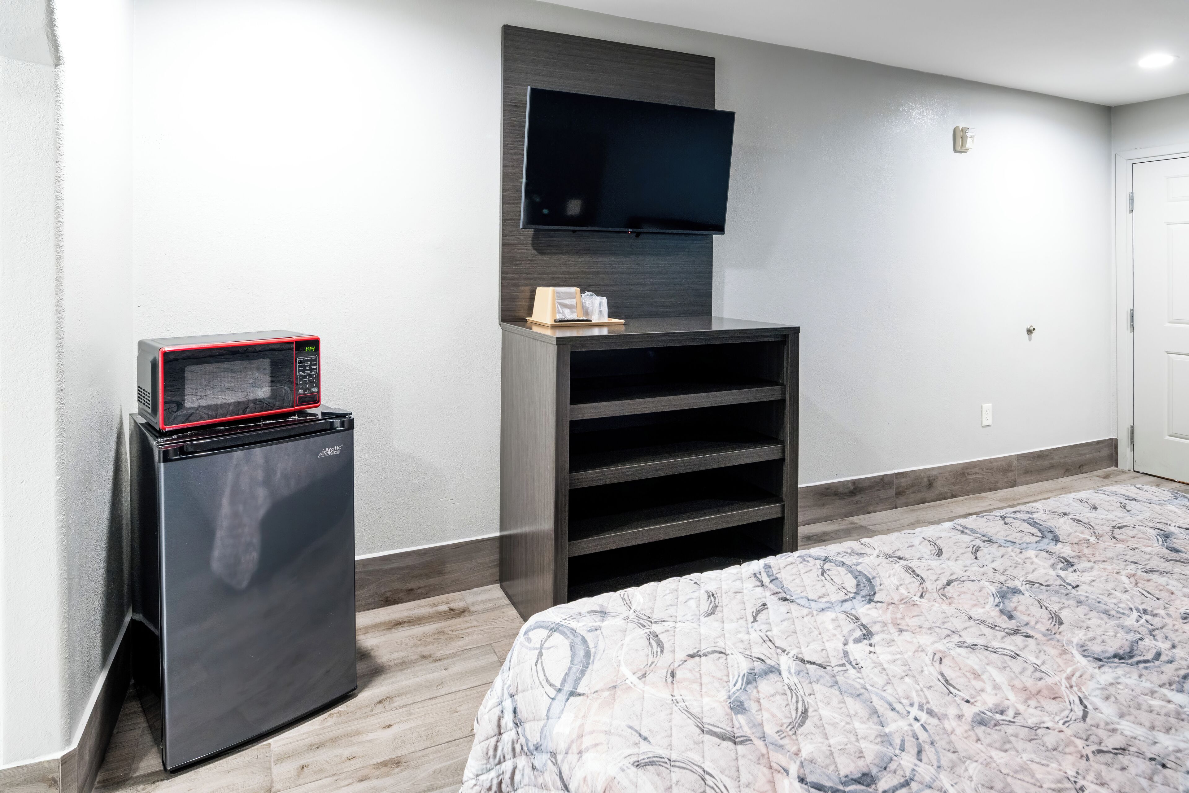 A compact room with a bed, a small refrigerator, a flat-screen TV mounted on the wall, and a built-in shelving unit.